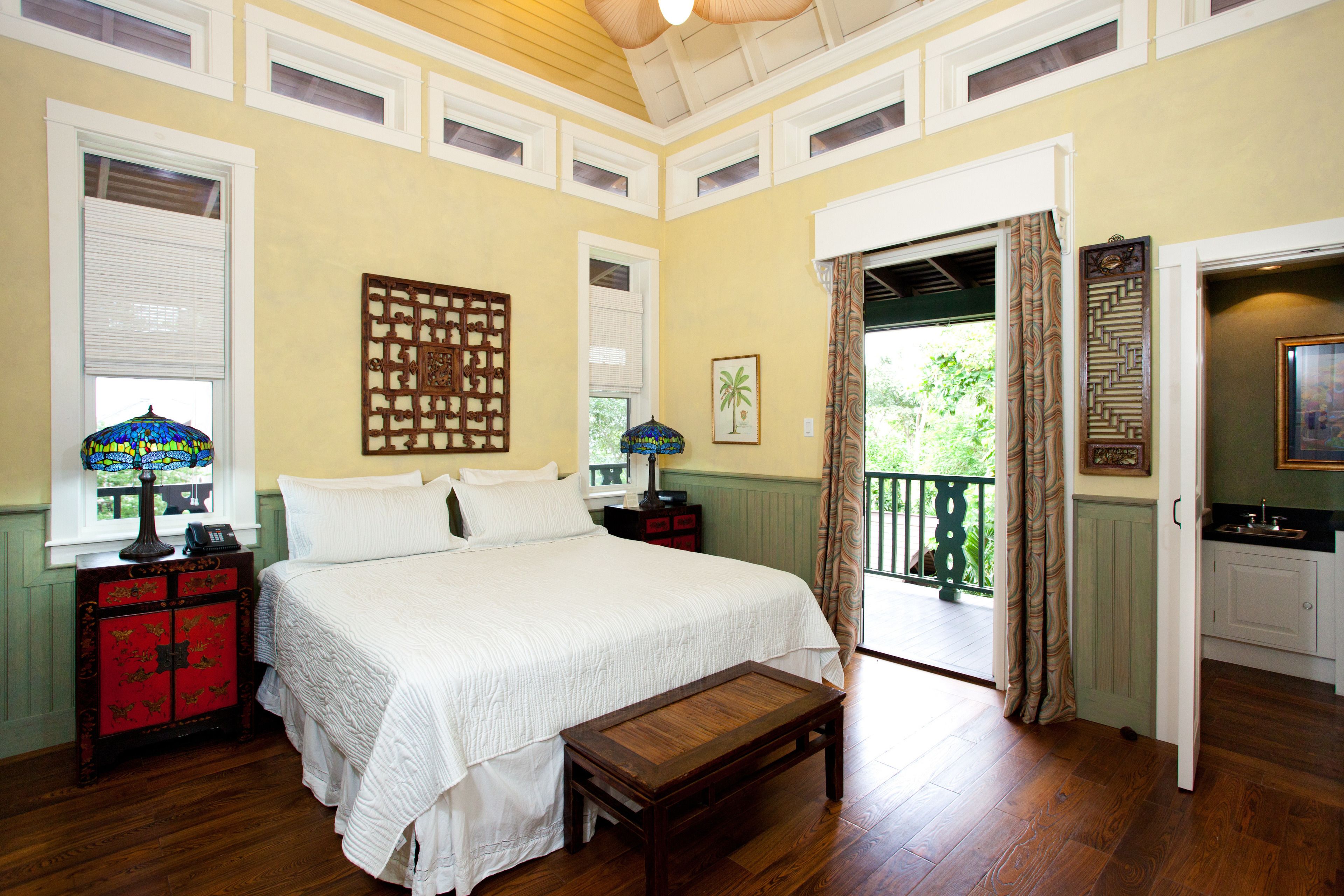 A bedroom with a large bed, a wooden bench, a nightstand, a lamp, and a view of the outdoors.