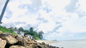 A beach with a rocky foreground, palm trees, and a few buildings in the background. -  The Relax Beach Resort (Ko Pha-ngan)