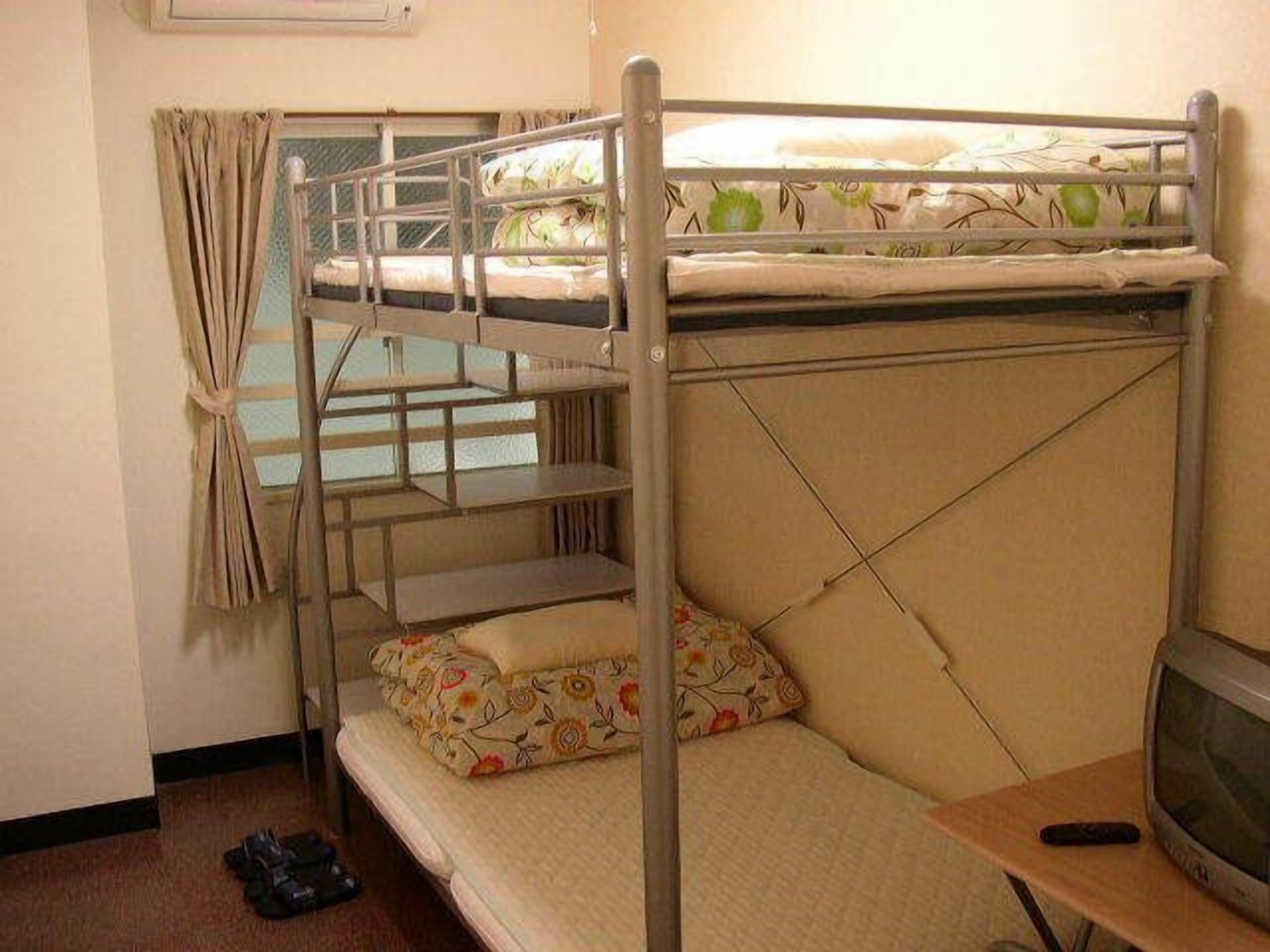 A bunk bed with a television on a wooden table, a window with curtains, and a pair of shoes on the floor.