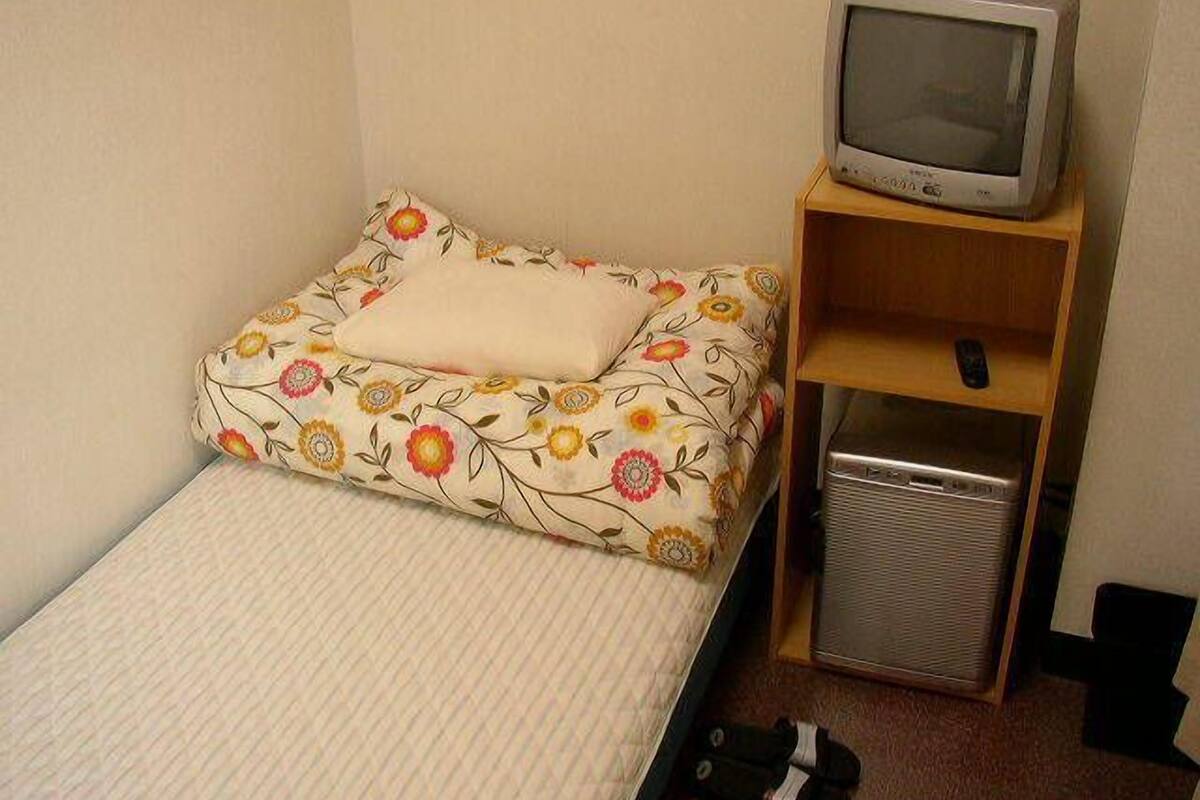 A small, single-bed room with a floral-patterned bedspread, a television on a wooden shelf, and a compact, metallic heater.