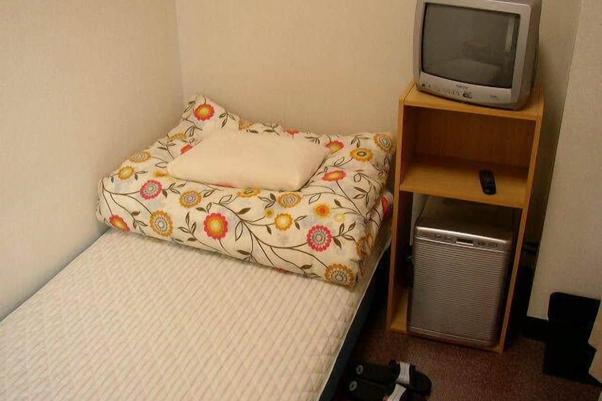 Quarto pequeno com uma cama de solteiro, colcha estampada com flores, televisão sobre uma prateleira de madeira e aquecedor metálico compacto.