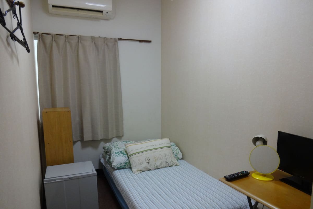 A small, neatly arranged bedroom with a bed, a desk with a computer and a mirror, a small heater, and a window with curtains.