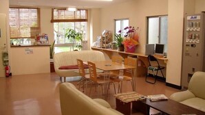 A spacious room with a dining area, a sofa, a small table, and a computer desk. There are windows with blinds, a fire extinguisher, and a vending machine. - Grace Naha - Hostel (Naha)