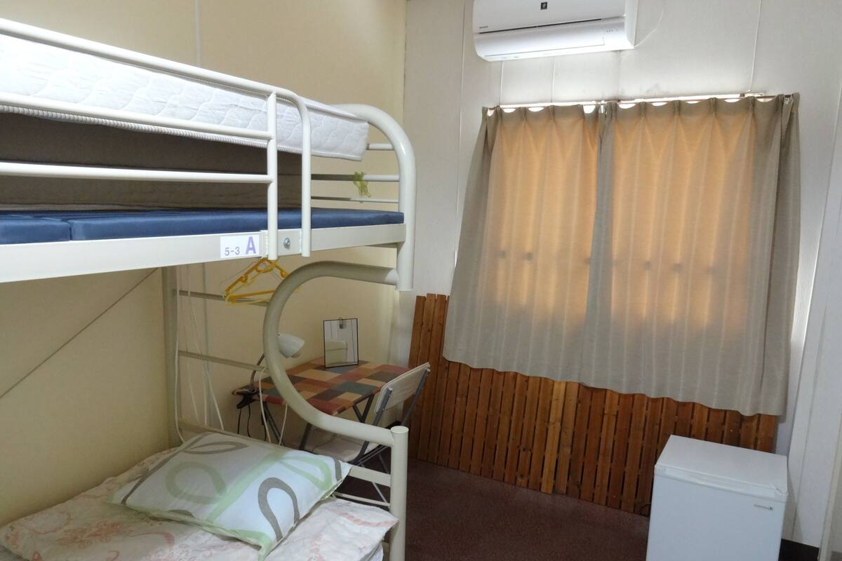 A bunk bed room with a desk, a chair, a small refrigerator, and a window with curtains.