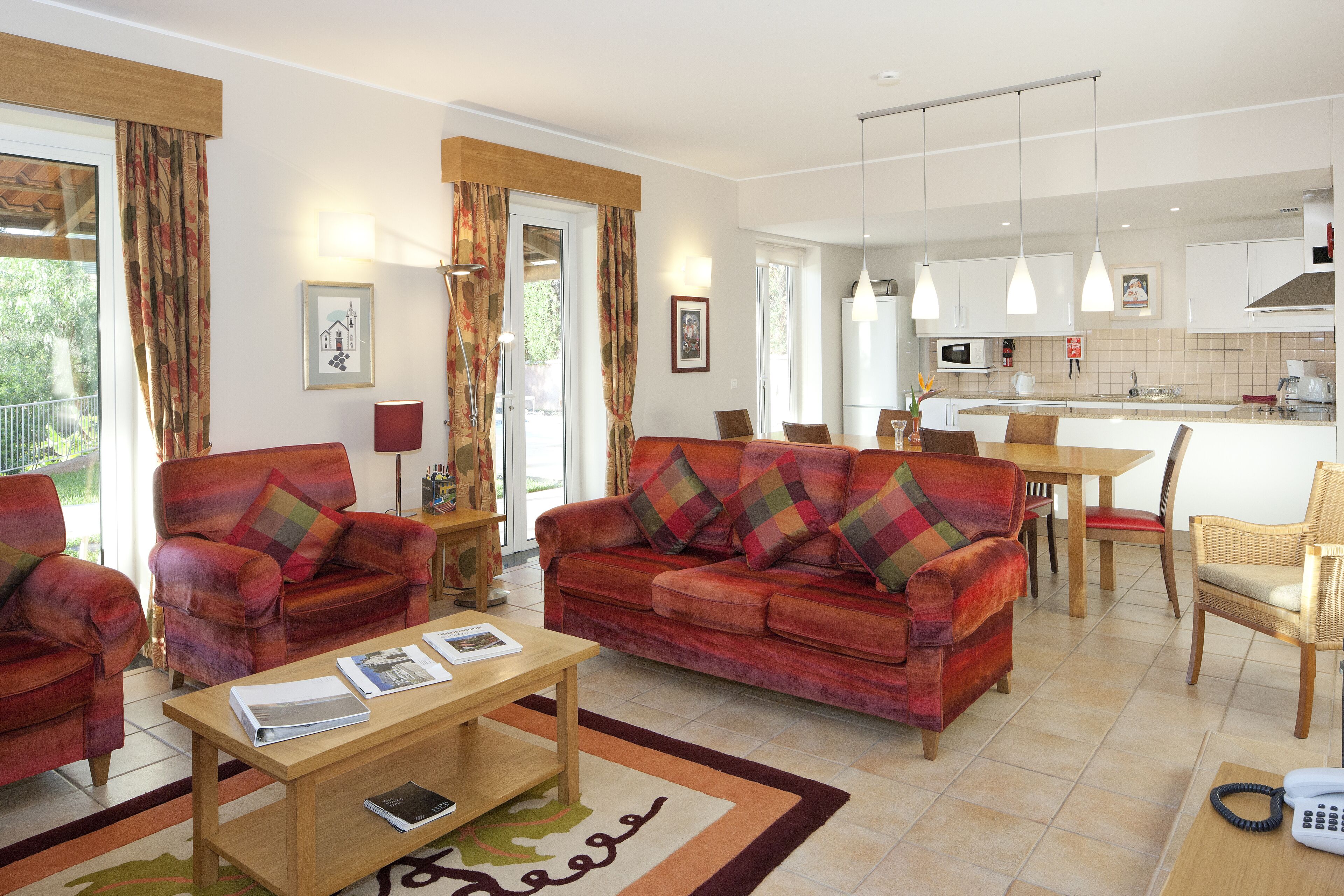 Una sala de estar moderna con un sofá, sillones y una mesa de centro. Hay un comedor con una mesa y sillas, y una cocina con gabinetes y electrodomésticos blancos.