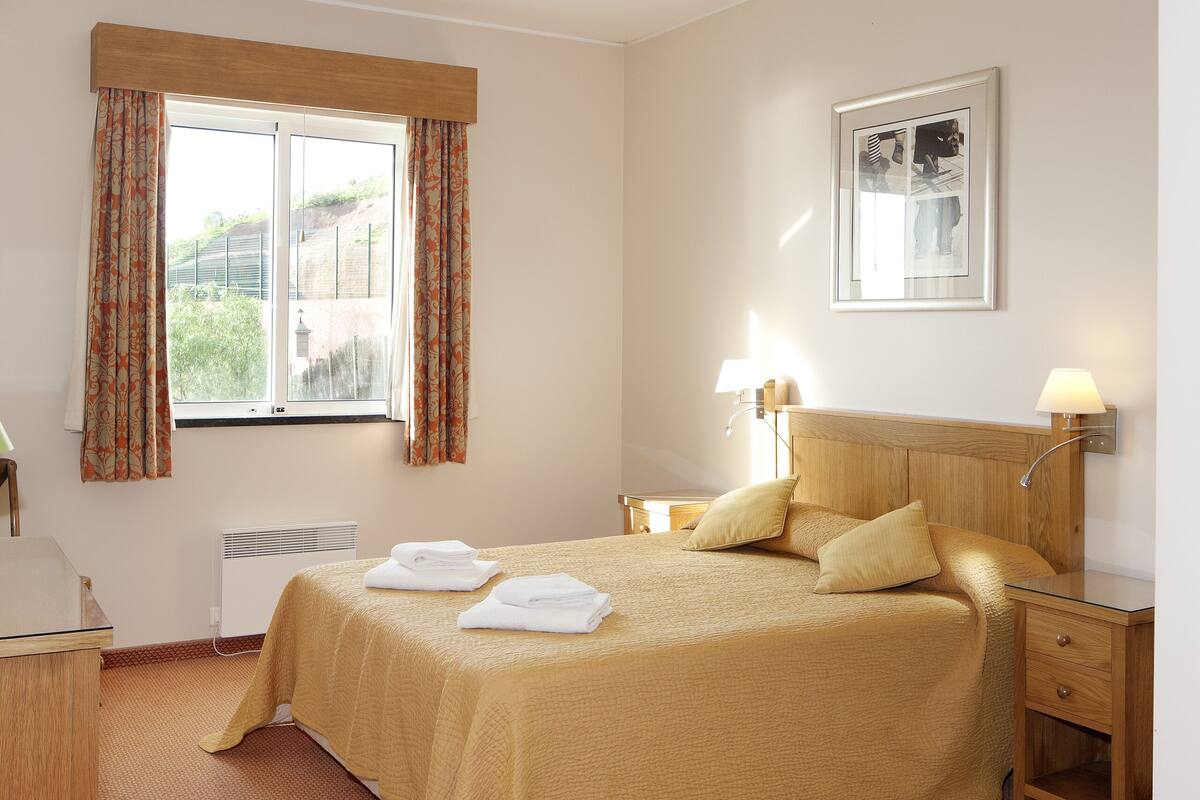 A hotel room with a bed, bedside lamps, a window with curtains, a framed picture on the wall, and a small bedside table.
