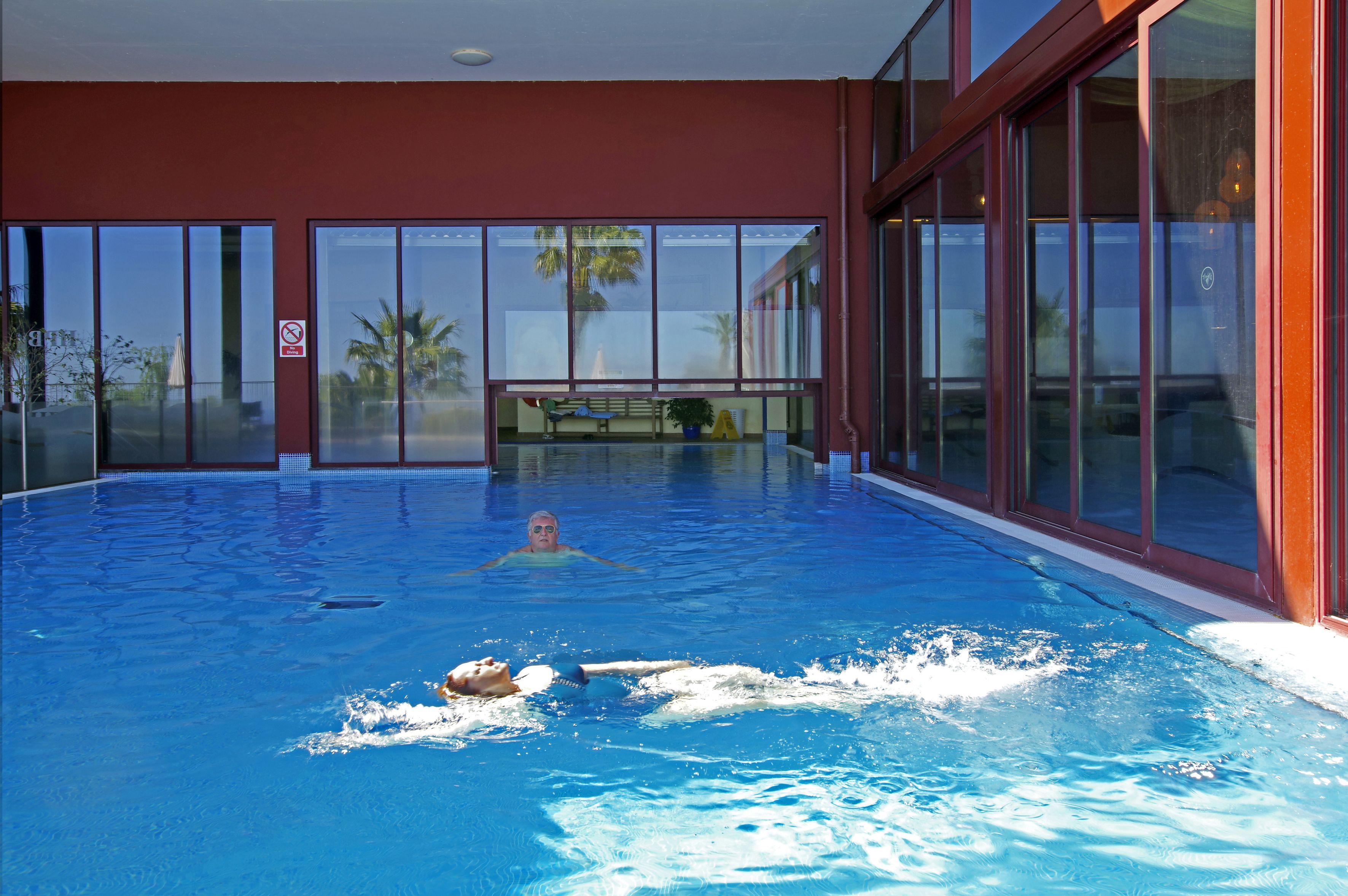 An indoor swimming pool with clear glass walls, a red exterior, and a bench in the background.