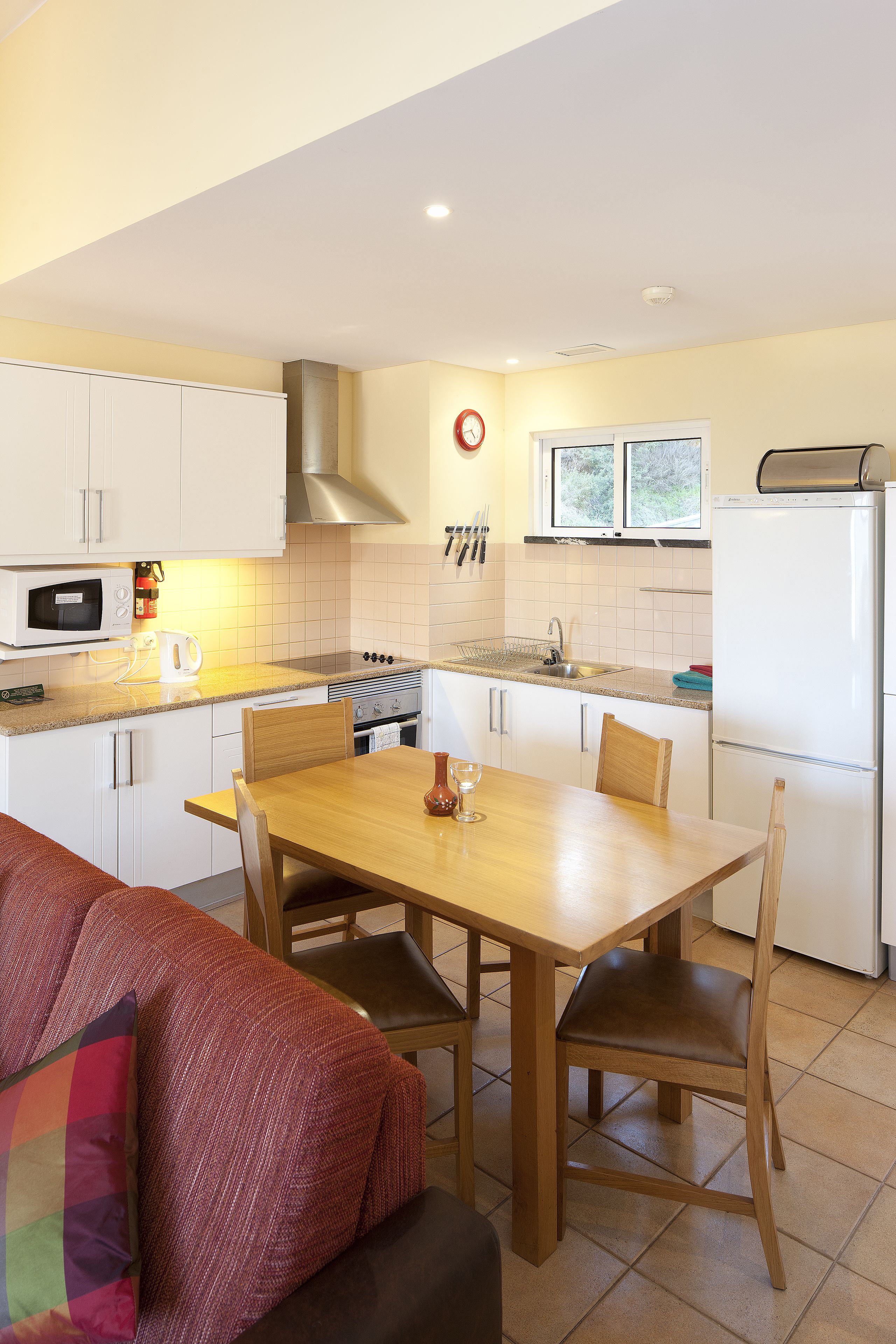 A compact kitchen with white cabinets, a microwave, stove, and a refrigerator. There is a dining table with chairs and a sofa with a colorful cushion.