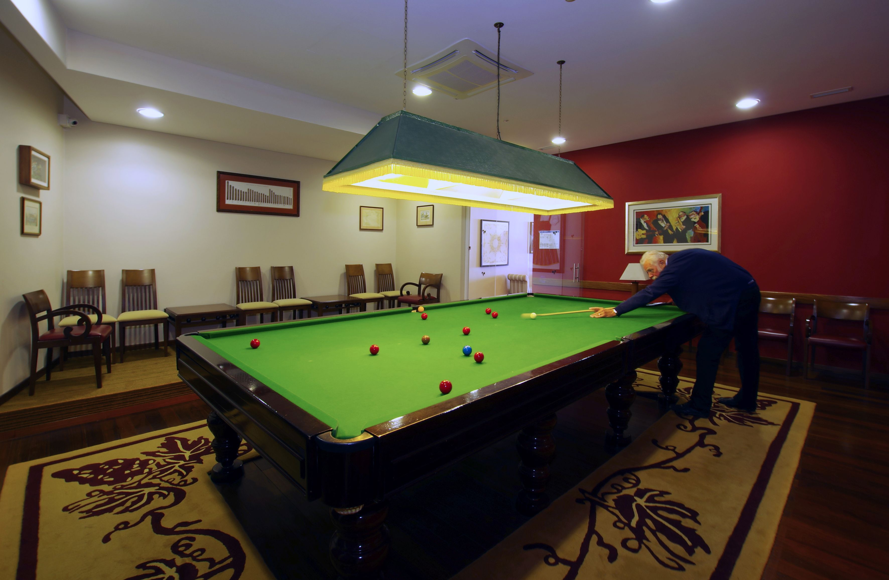 A pool table with balls in a room with red walls, a green overhead light, and several chairs.