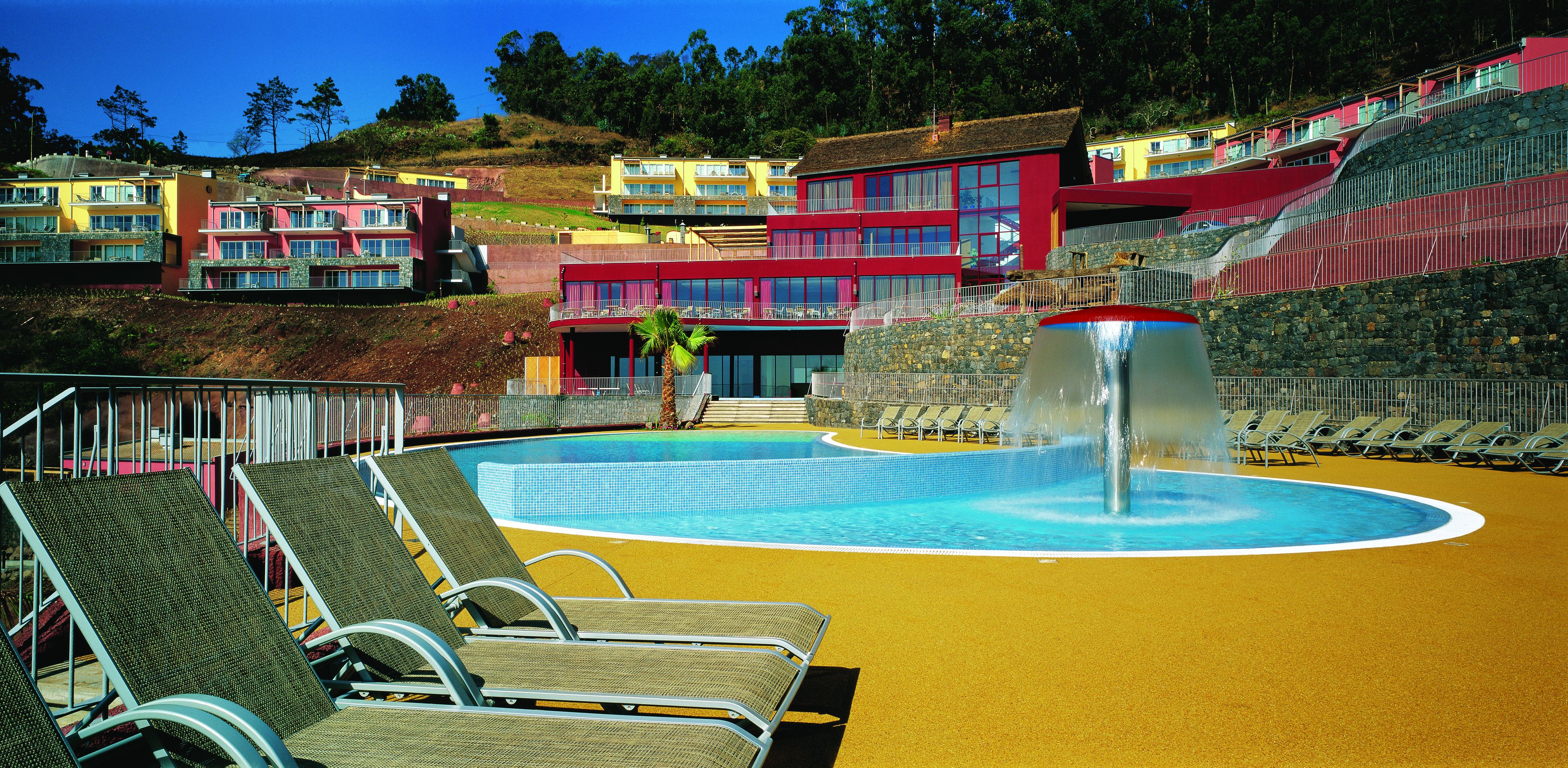 A pool area with lounge chairs, a fountain, and colorful buildings in the background.