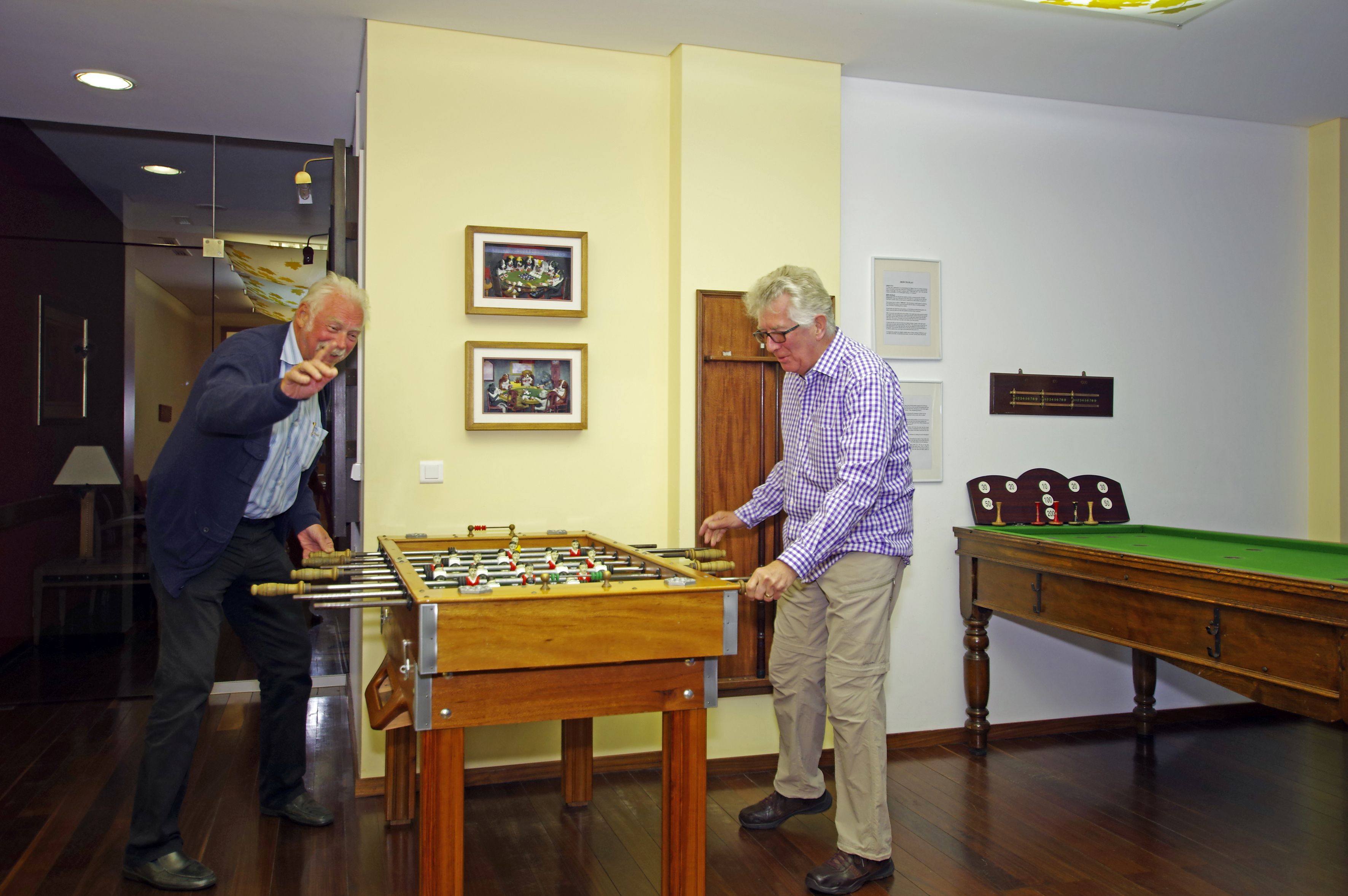 Deux hommes jouent au baby-foot dans une pièce où se trouvent une table de billard et des tableaux encadrés accrochés au mur.