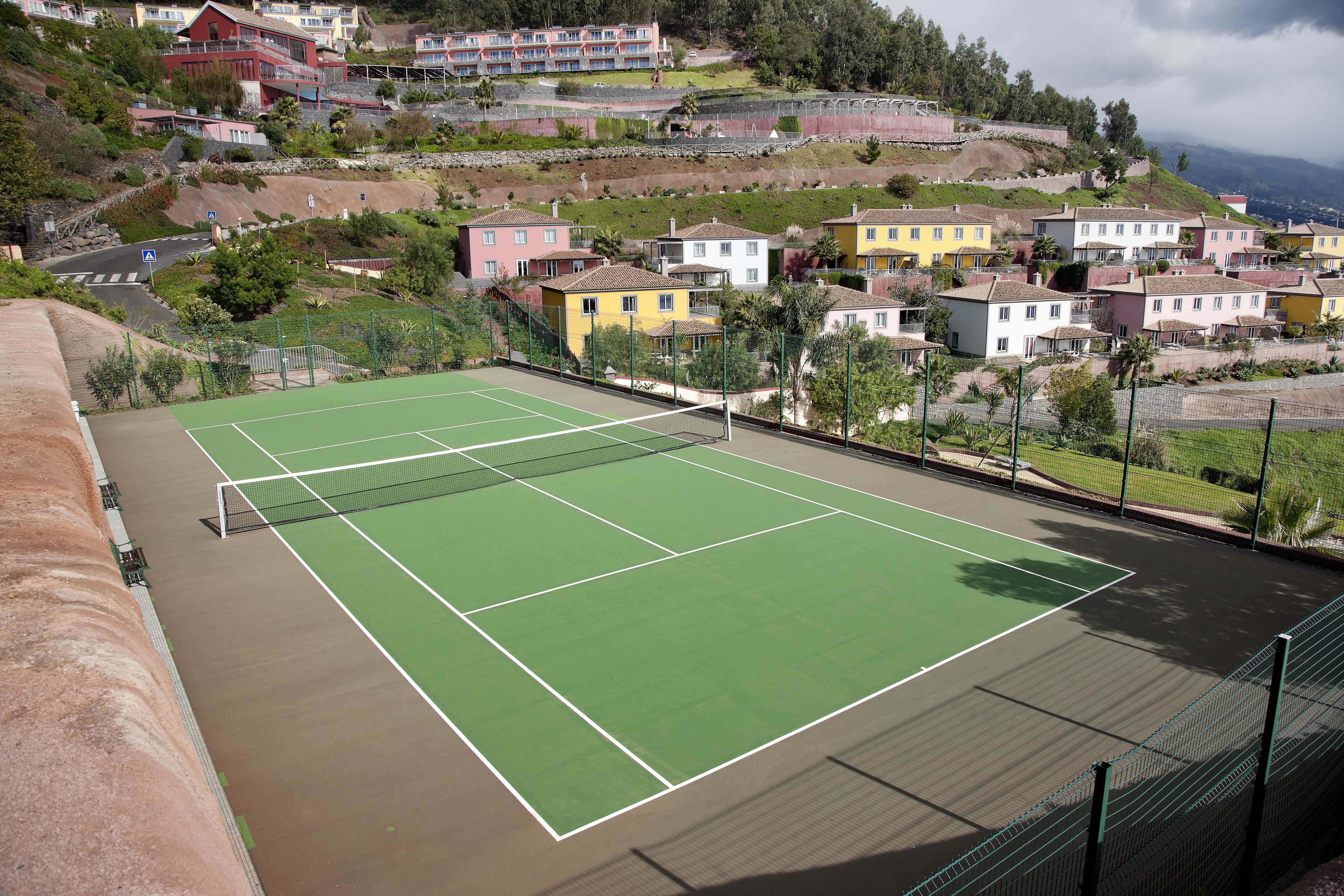 A tennis court with a net, surrounded by a fence and a hillside with colorful houses.