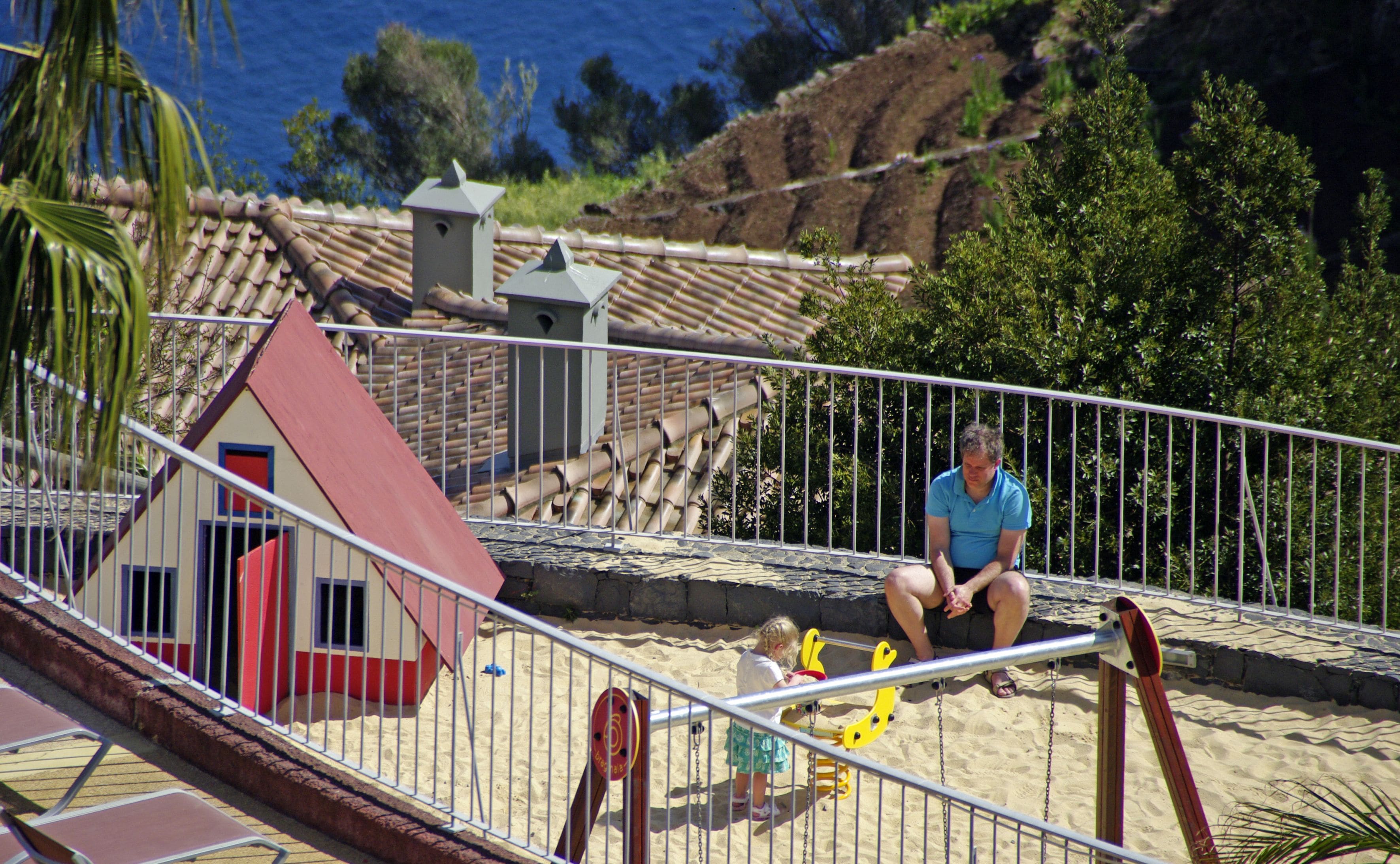 A beach scene with a small house, a man sitting on a swing, and a child playing in the sand.