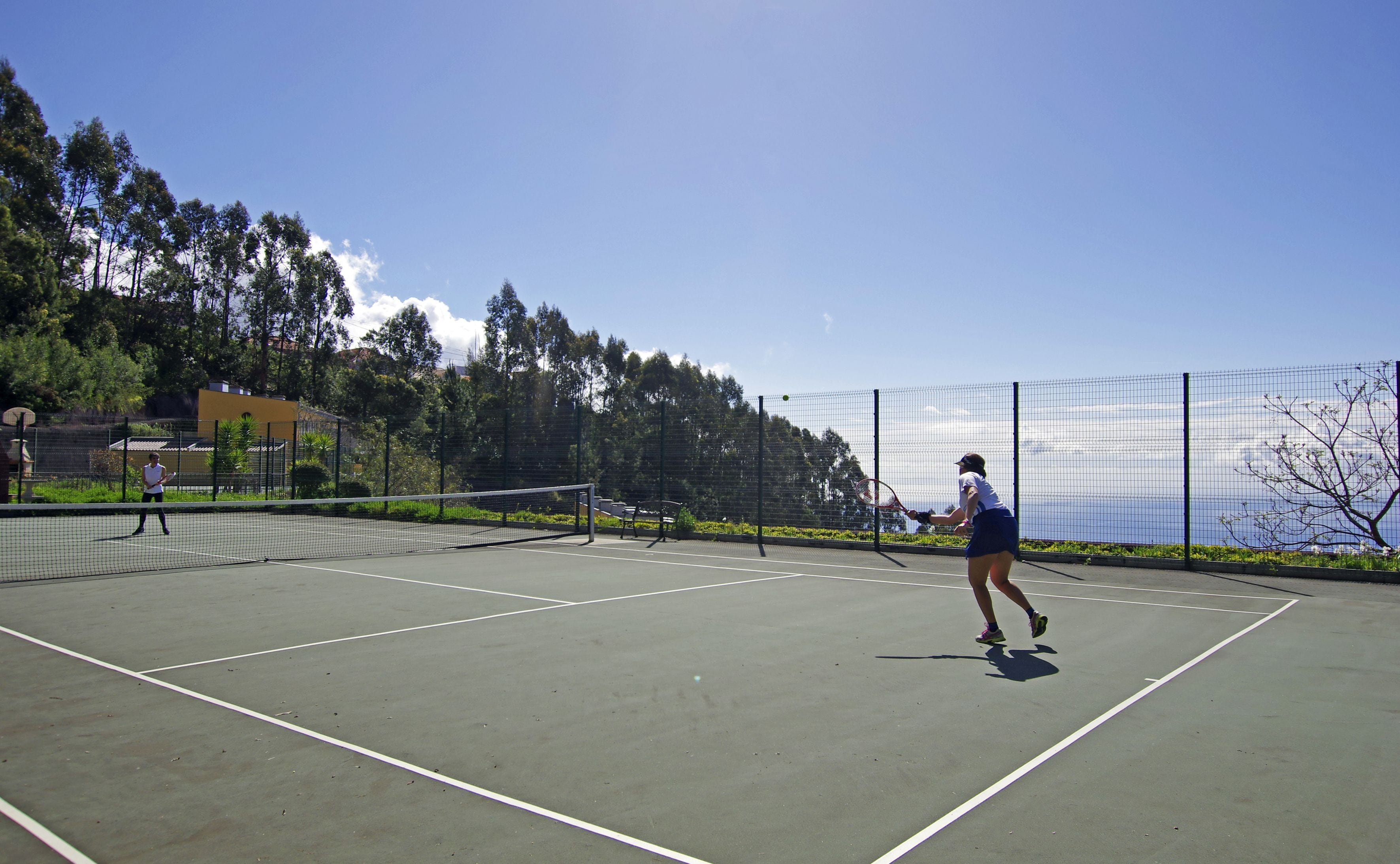 An outdoor tennis court with a net, surrounded by a fence and trees.