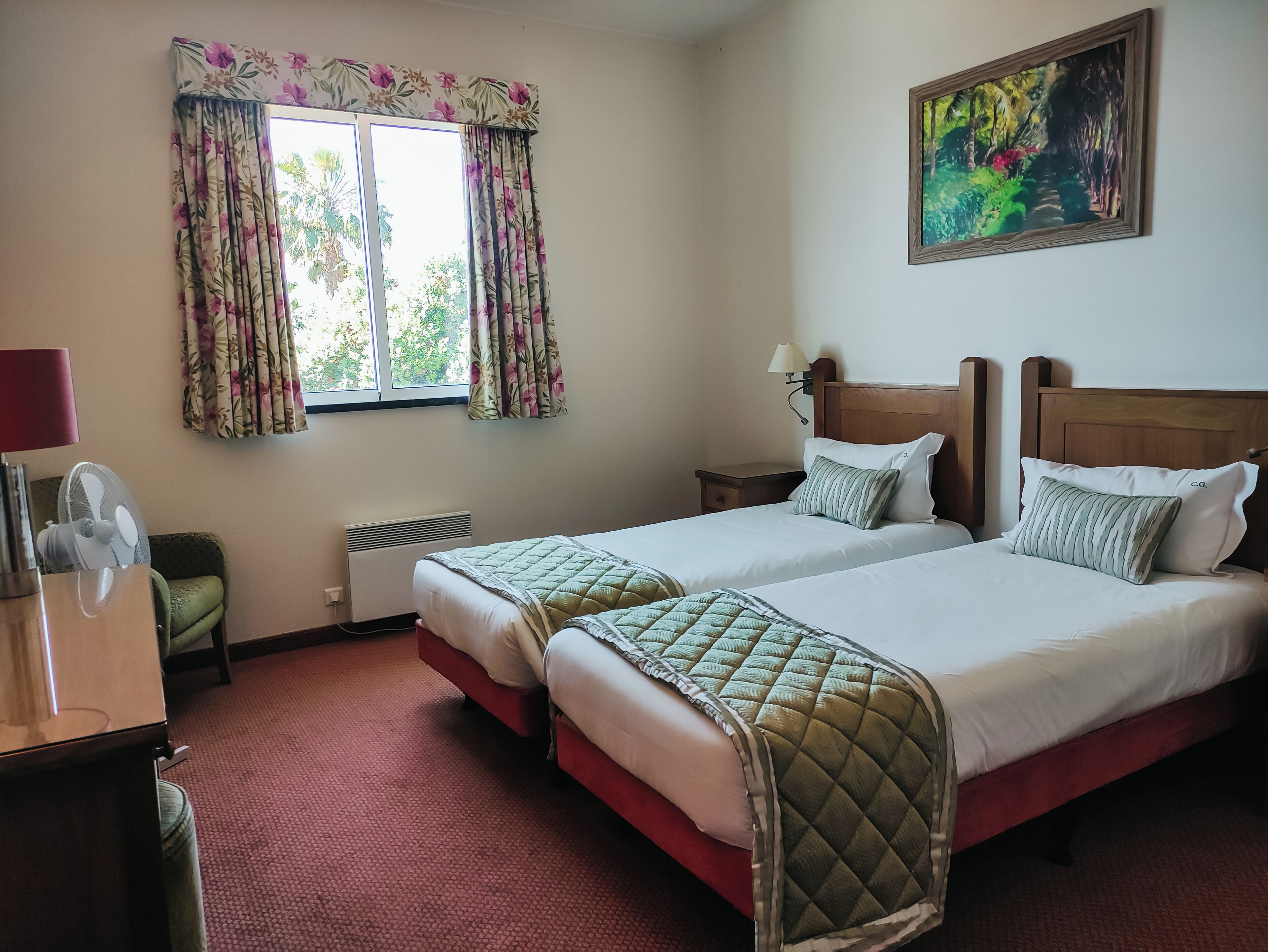 A hotel room with two beds, a desk, a chair, a radiator, and a window with curtains.