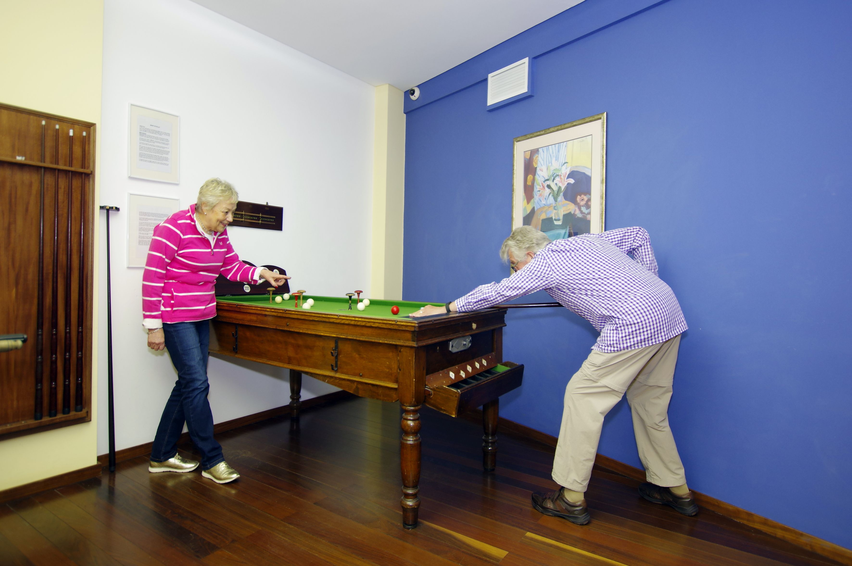 Two people playing pool in a room with wooden flooring and a blue wall.