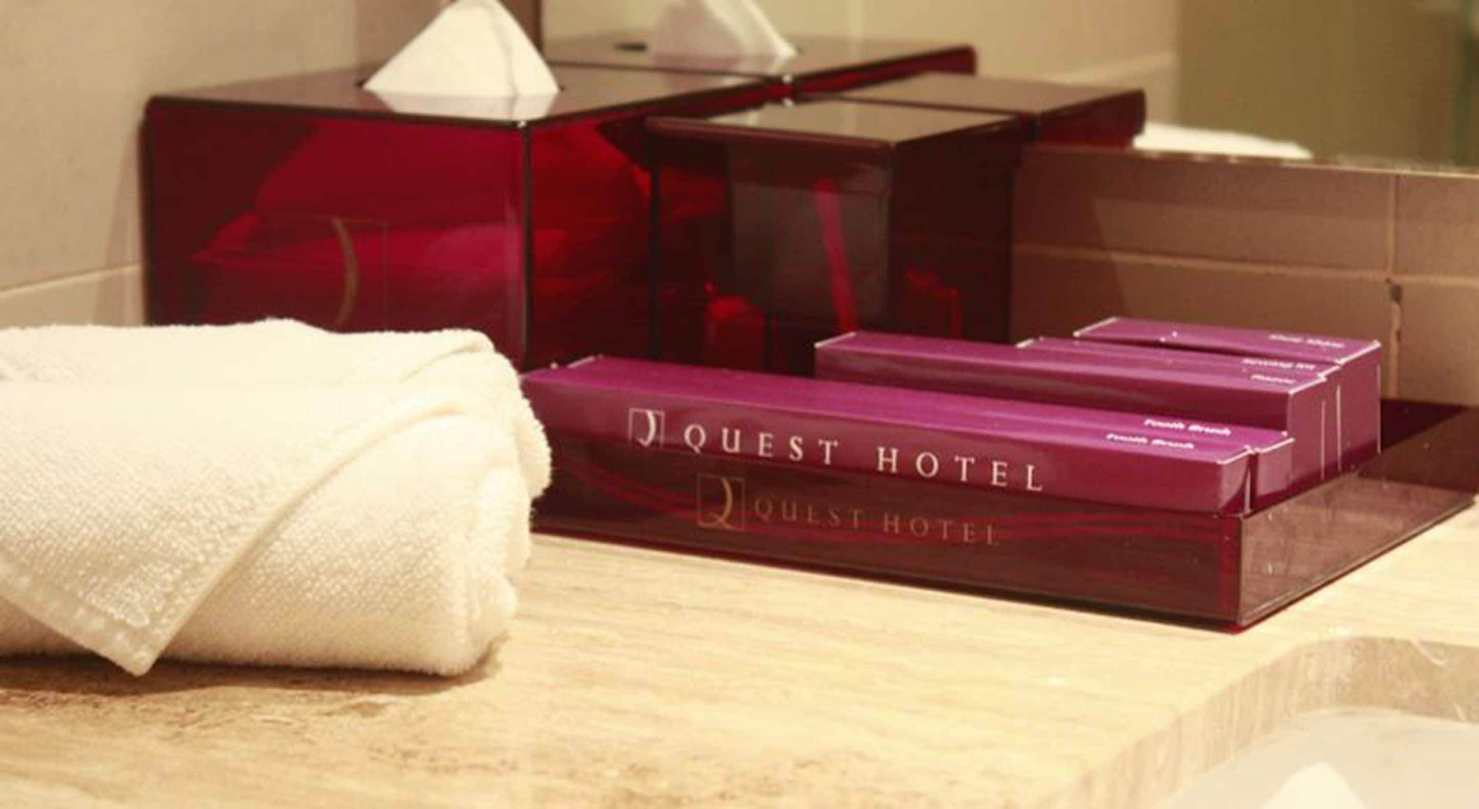Une serviette blanche et deux boîtes violettes portant l’inscription « Quest Hotel » sont posées sur une surface en bois.