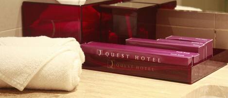 Une serviette blanche et deux boîtes violettes portant l’inscription « Quest Hotel » sont posées sur une surface en bois.