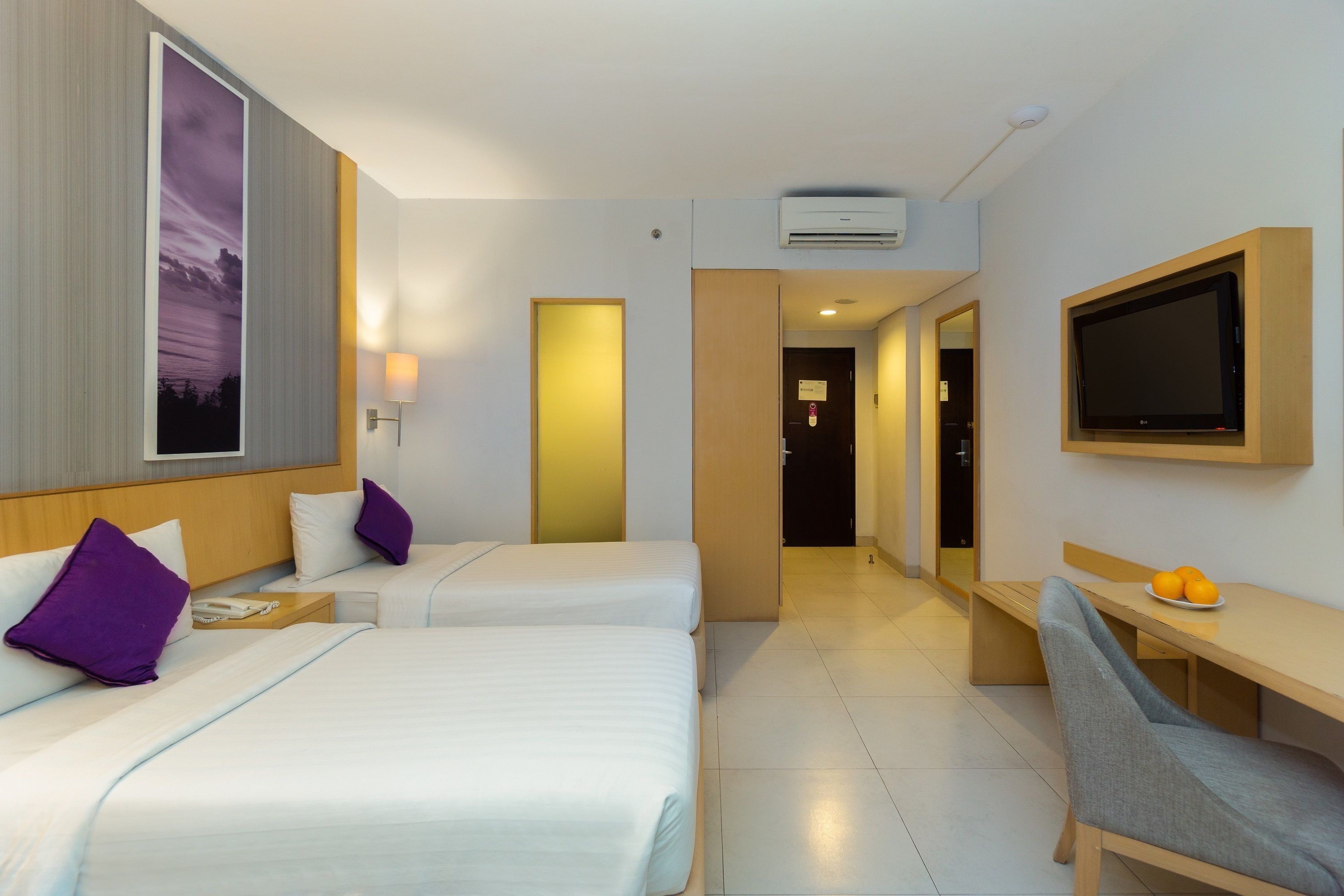 A hotel room with two beds, a desk, a chair, a TV, and a wall-mounted air conditioner.