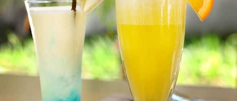 Two drinks in glasses, one with a blue layer and the other with a yellow layer, and a slice of orange on the right glass.