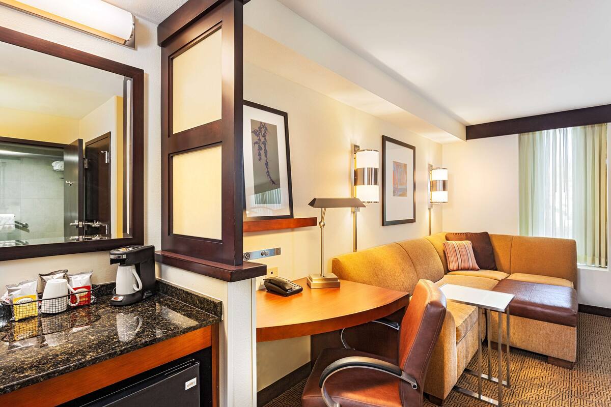 A hotel room with a kitchenette, a sofa, a desk, and a large window.