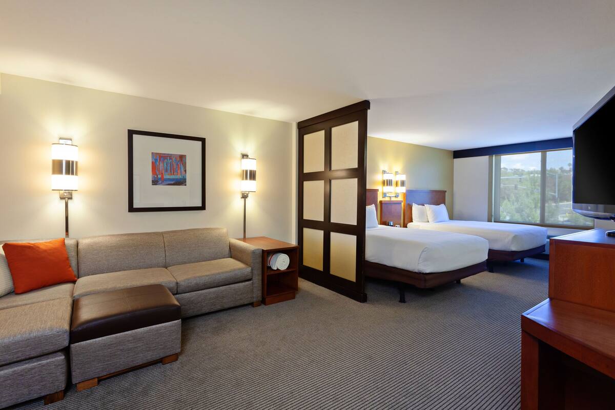 A hotel room with a sofa, two beds, a TV, and wall-mounted lamps.