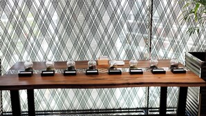Une table en bois, sur laquelle sont disposés des bocaux en verre et des boîtes noires, le tout placé devant une clôture en métal à motifs.