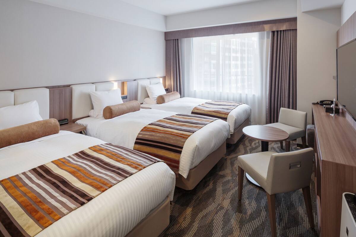 A hotel room with three beds, a desk, a chair, and a large window with curtains.