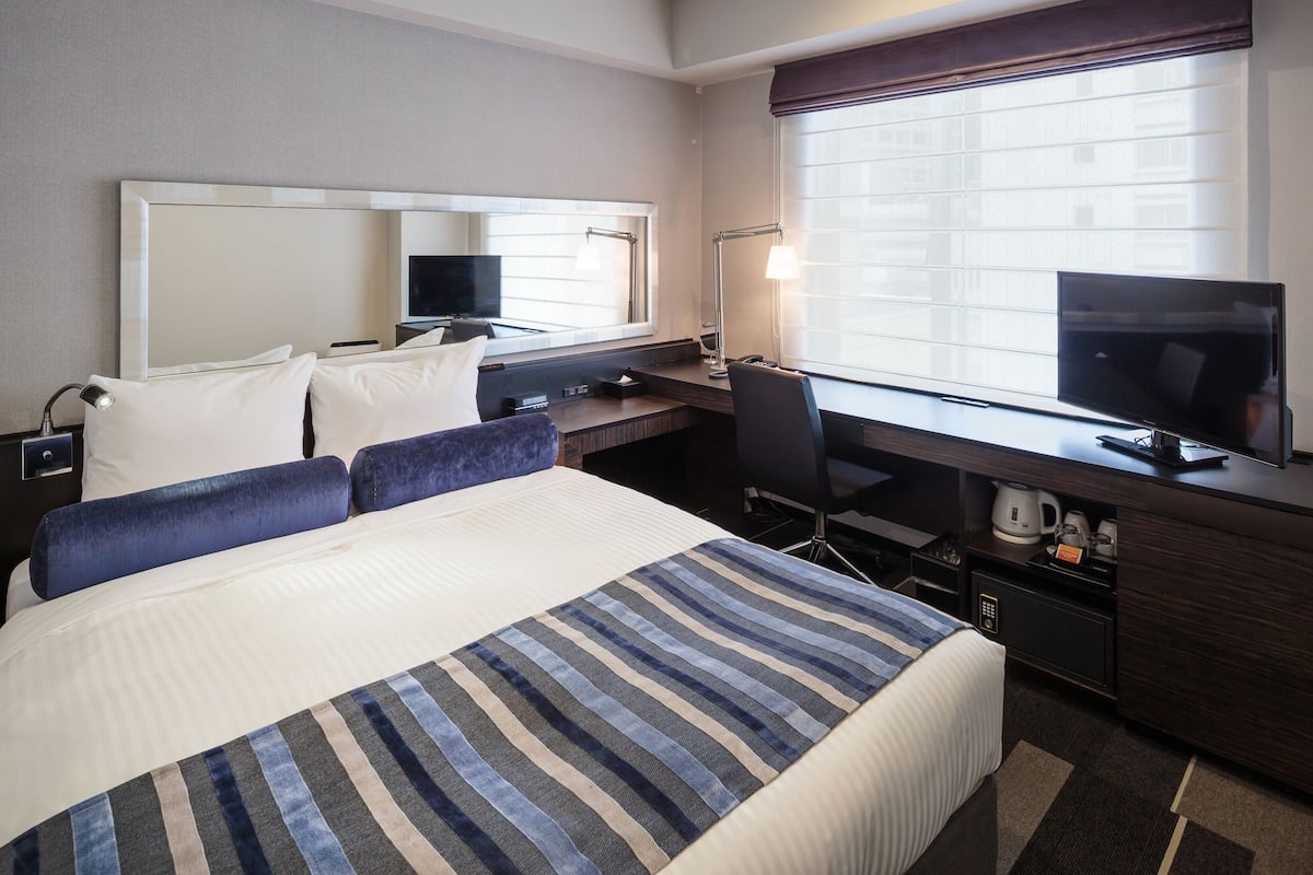A hotel room with a large bed, a desk with a computer, a TV, and a window with blinds.