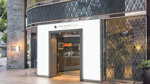 Une entrée d’hôtel moderne avec une porte en verre, une enseigne indiquant « HÔTEL MYSTAYS Doha Premier », et une clôture métallique ornée d’un motif géométrique.