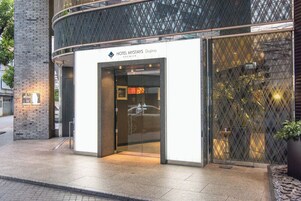 A modern hotel entrance with a glass door, a sign reading "HOTEL MYSTAYS Doha Premier," and a metal fence with a geometric pattern.