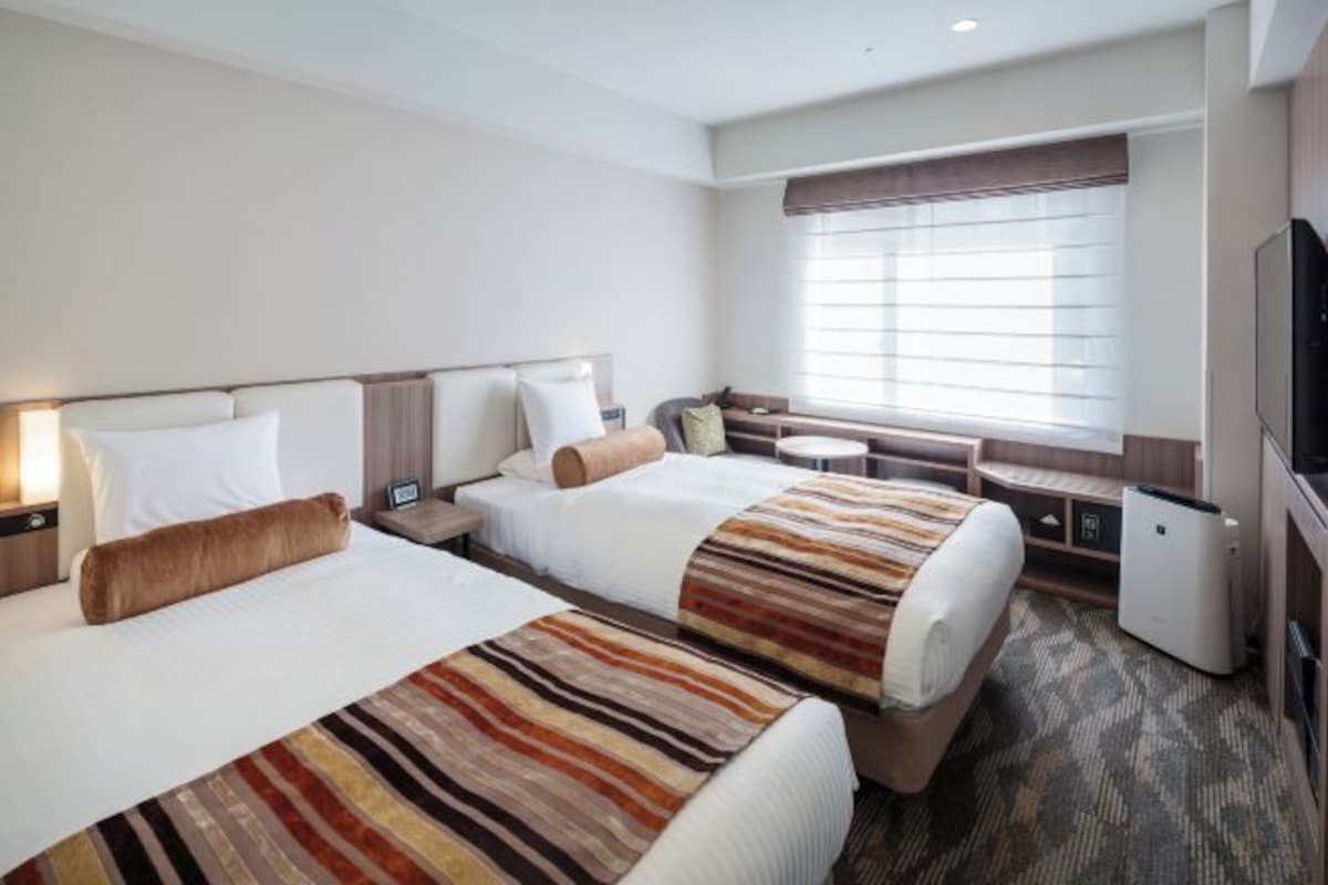A hotel room with two beds, a TV, a desk, and a window with blinds.