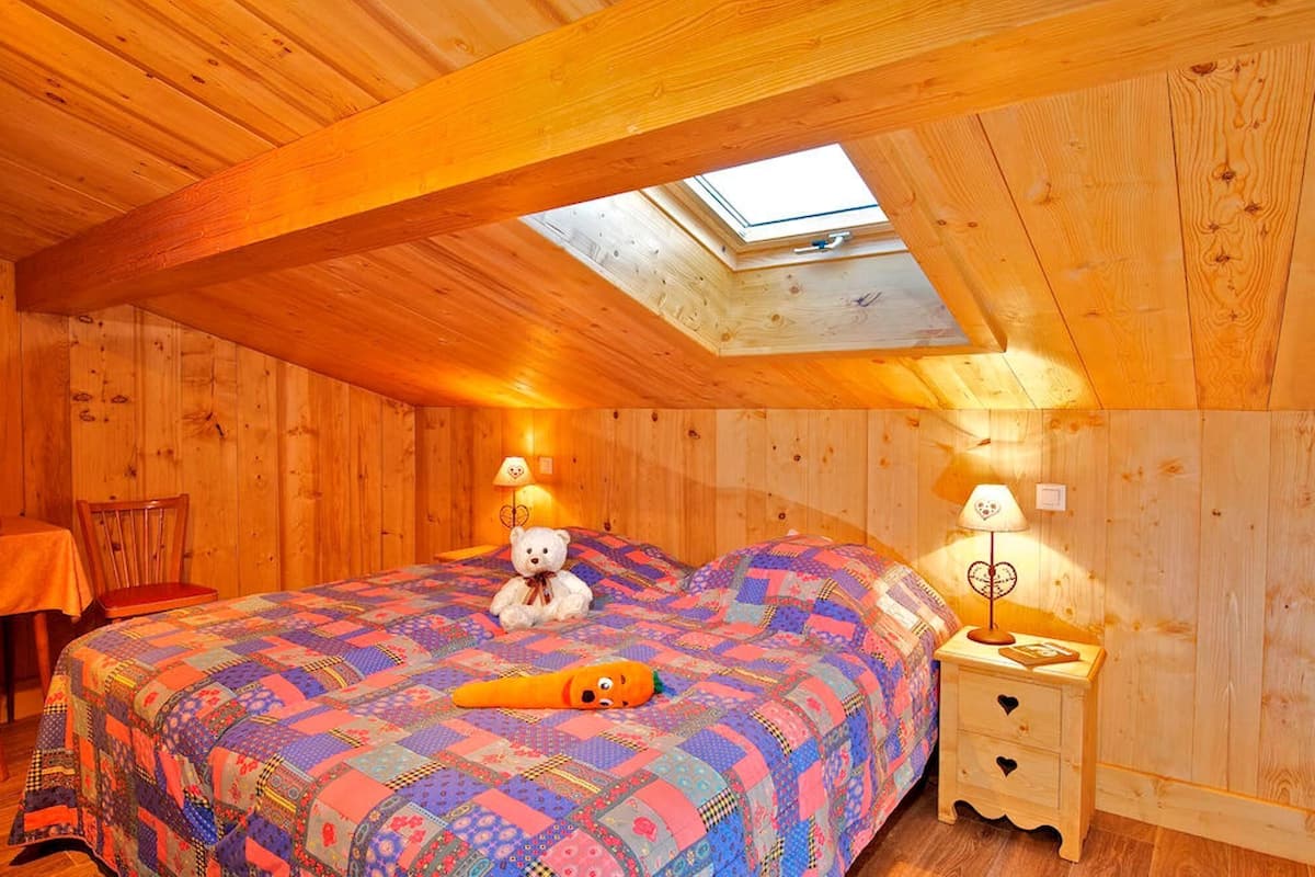 Un dormitorio acogedor en el ático con un tragaluz, una cama con un colcha colorida, un osito de peluche y una mesita de noche con una lámpara.