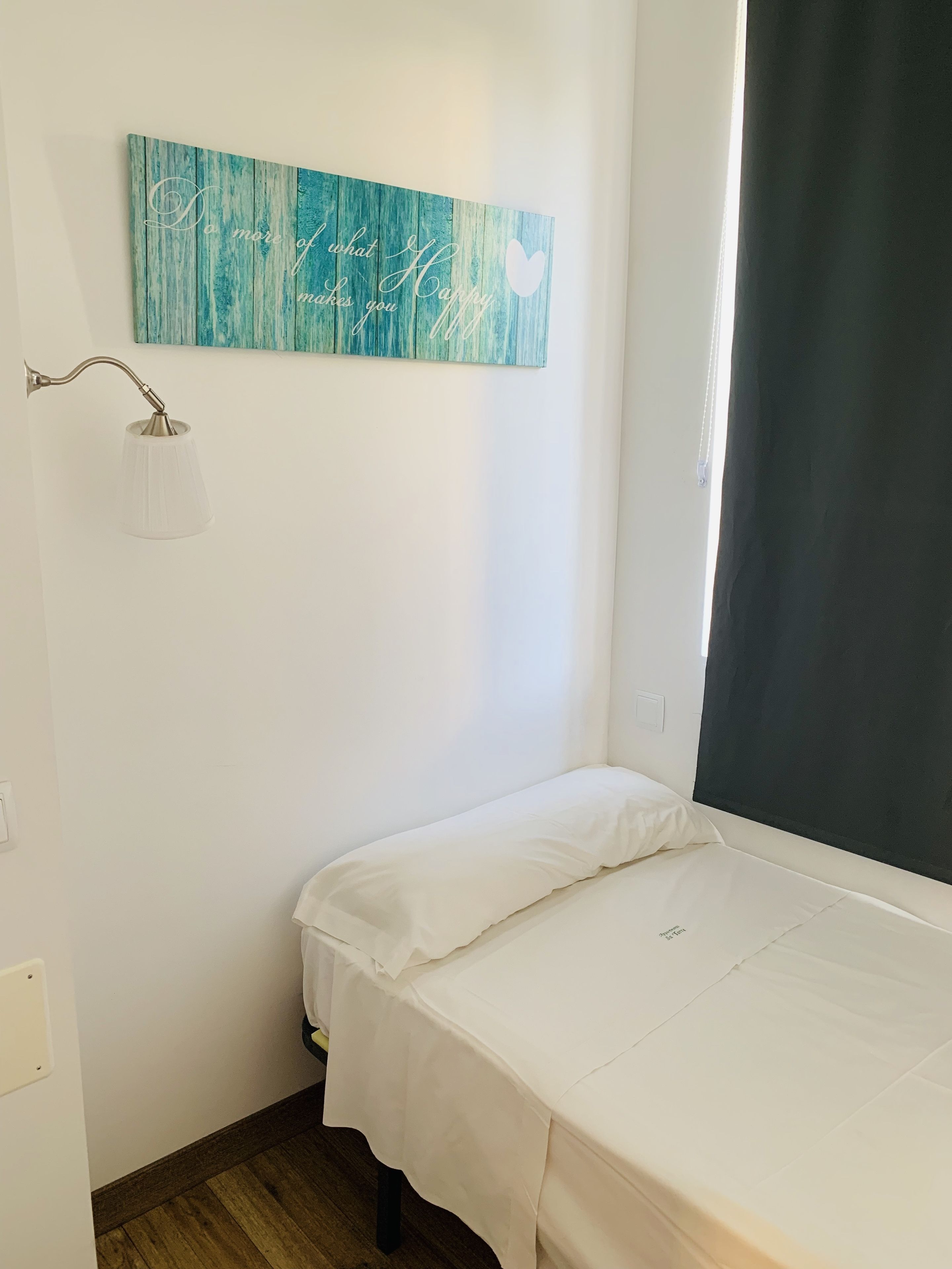 A small, neatly made bed with white bedding in a room with a teal wall art piece and a white lamp.