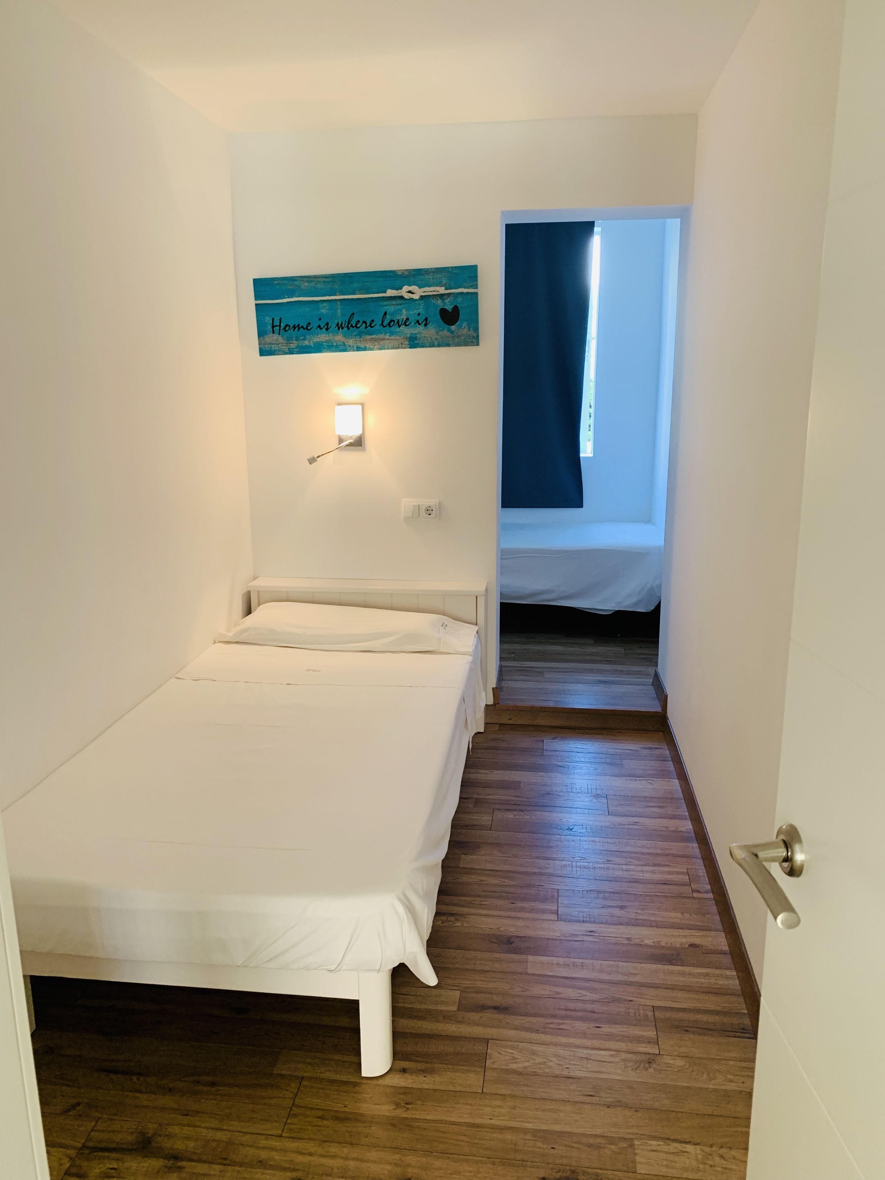 A narrow room with a single bed, a small wall-mounted light, and a blue sign with text.