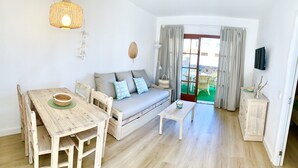 Een gezellige woonkamer met een houten eettafel, een bank met kussens, een klein bijzettafeltje en een rieten lamp.
