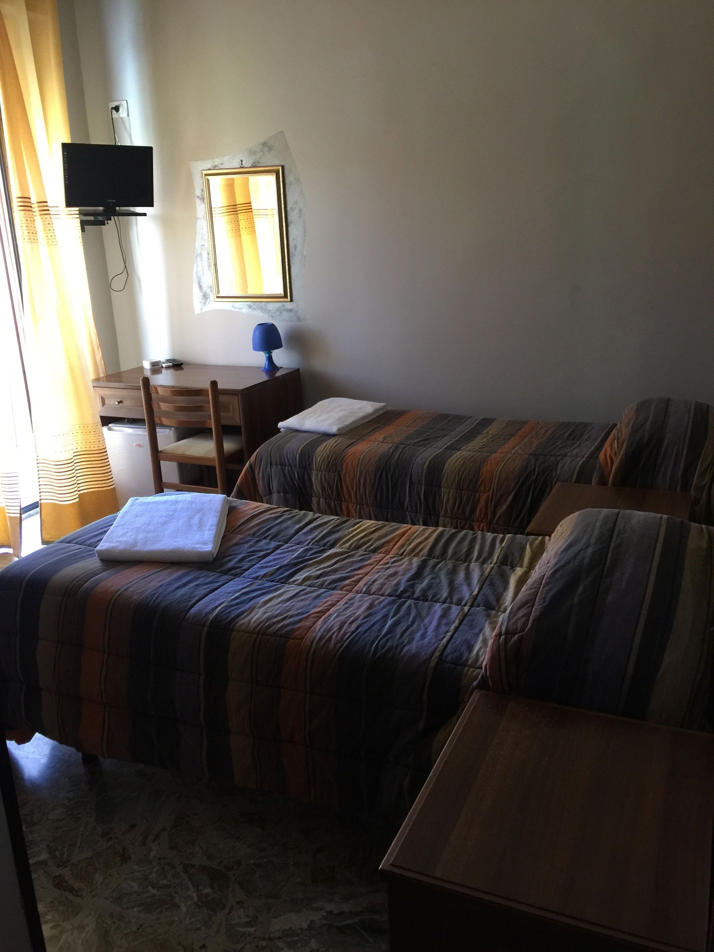 Una stanza con due letti, una scrivania, una sedia, una TV e una finestra con tende.