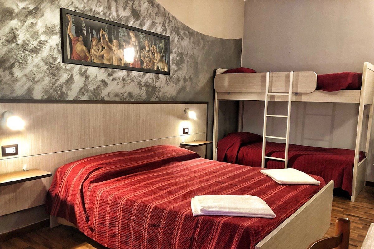 A room with a bunk bed, a single bed with a red blanket, a wooden nightstand, a framed picture on the wall, and a wooden floor.