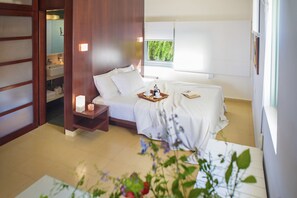A hotel room with a bed, bedside table, and a window with a view of greenery. - Nereids (Sitia)