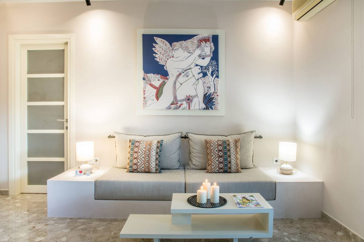 A modern living room with a sofa, two side tables with lamps, a coffee table with candles, and a wall-mounted artwork.