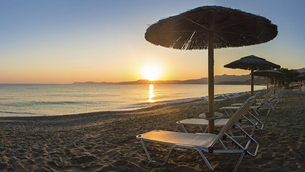 A beach with rows of sun loungers and thatched umbrellas, a clear view of the sunset over the sea, and a distant cityscape. - Nereids (Sitia)