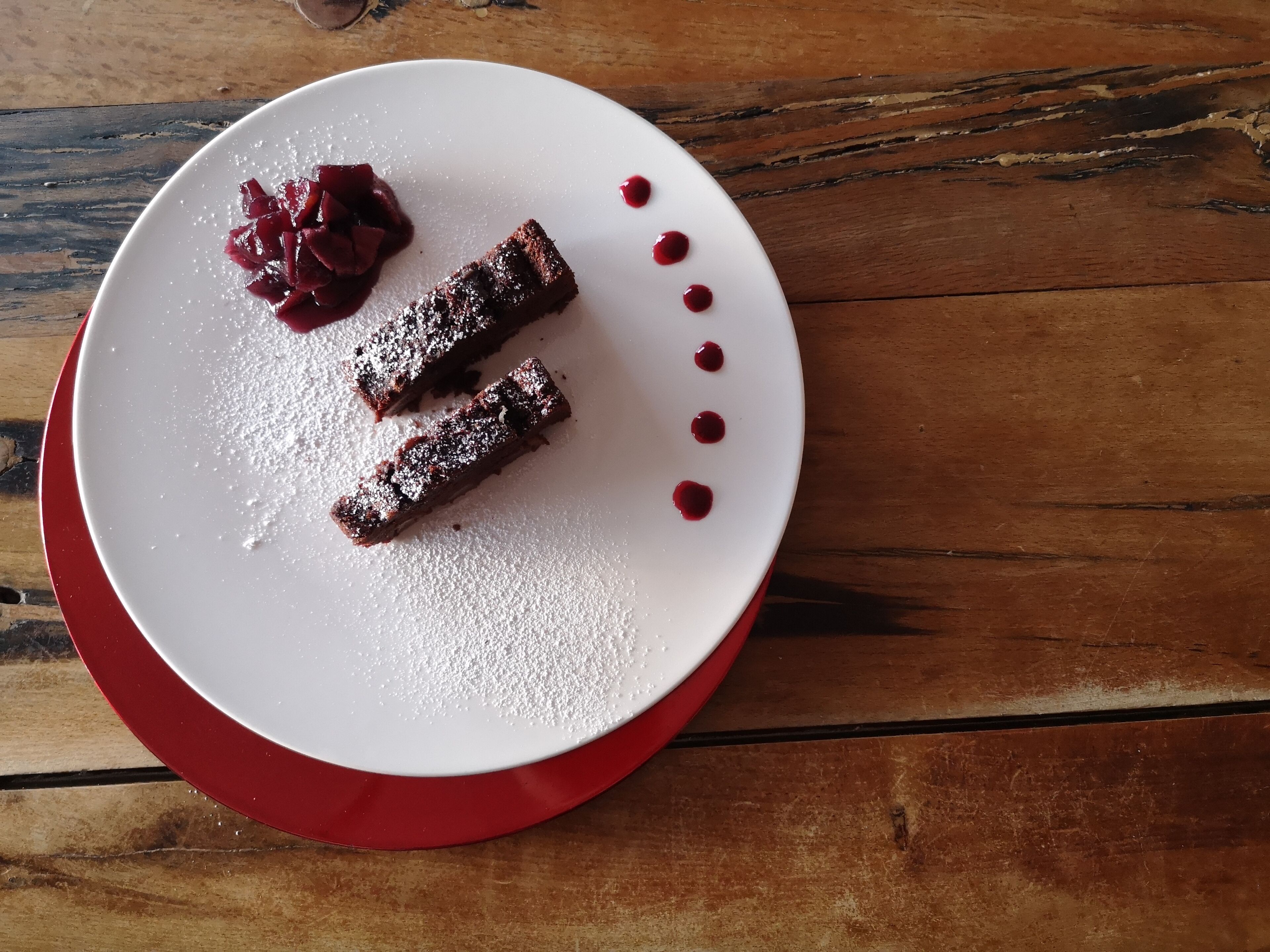 Un plato de postre con dos brownies y un puré de frutos rojos sobre una mesa de madera.