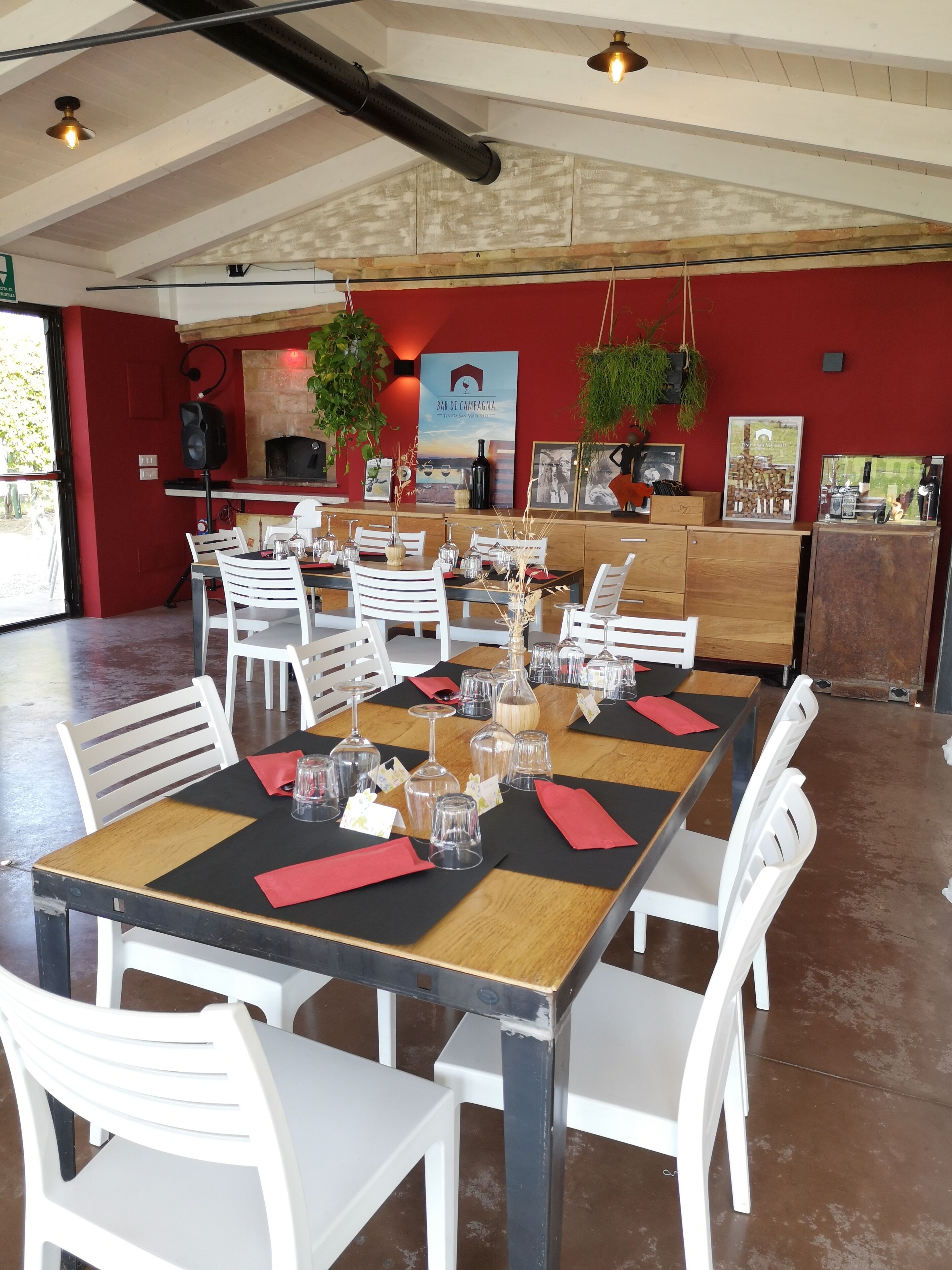 A restaurant with a long wooden table set for dining, white chairs, and a red wall with a poster.