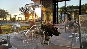 Una mesa lista para comer, con cristalería transparente y un florero con flores, con un jardín y un edificio como fondo.