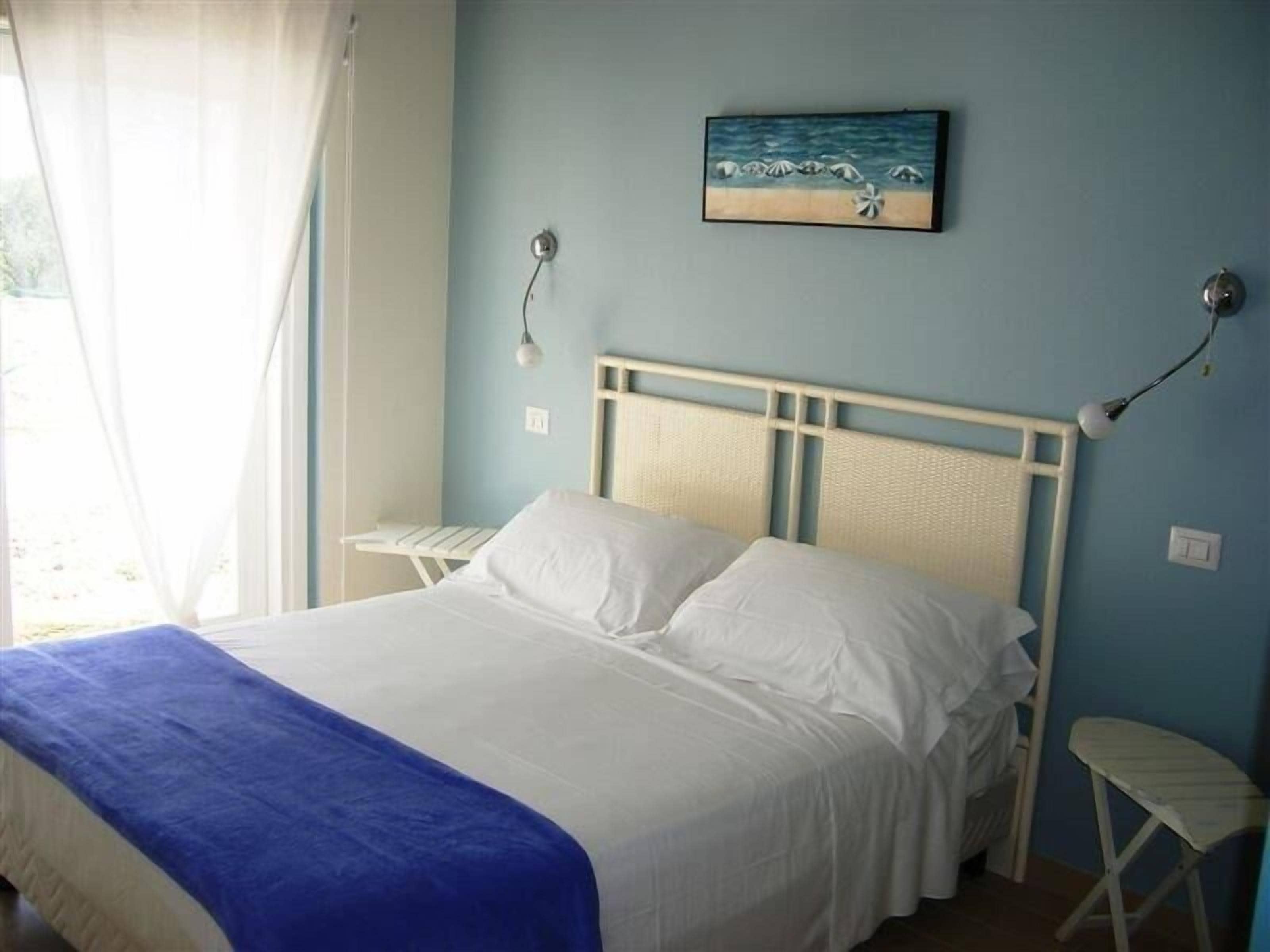 A bedroom with a bed, a nightstand, a wall-mounted lamp, and a painting of a beach scene.