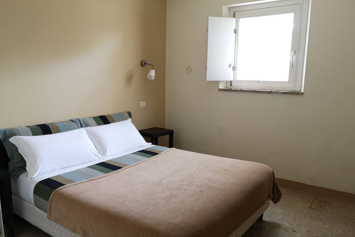 A bed with a striped headboard, a white pillow, and a beige blanket. There is a small table beside the bed and a window with an open door.