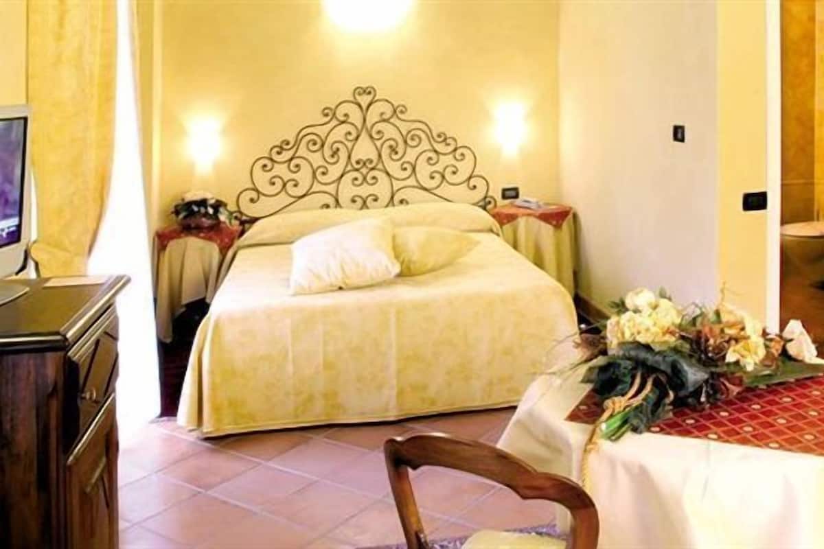 A hotel room with a bed, a desk with a computer, a chair, and a table with a floral arrangement.