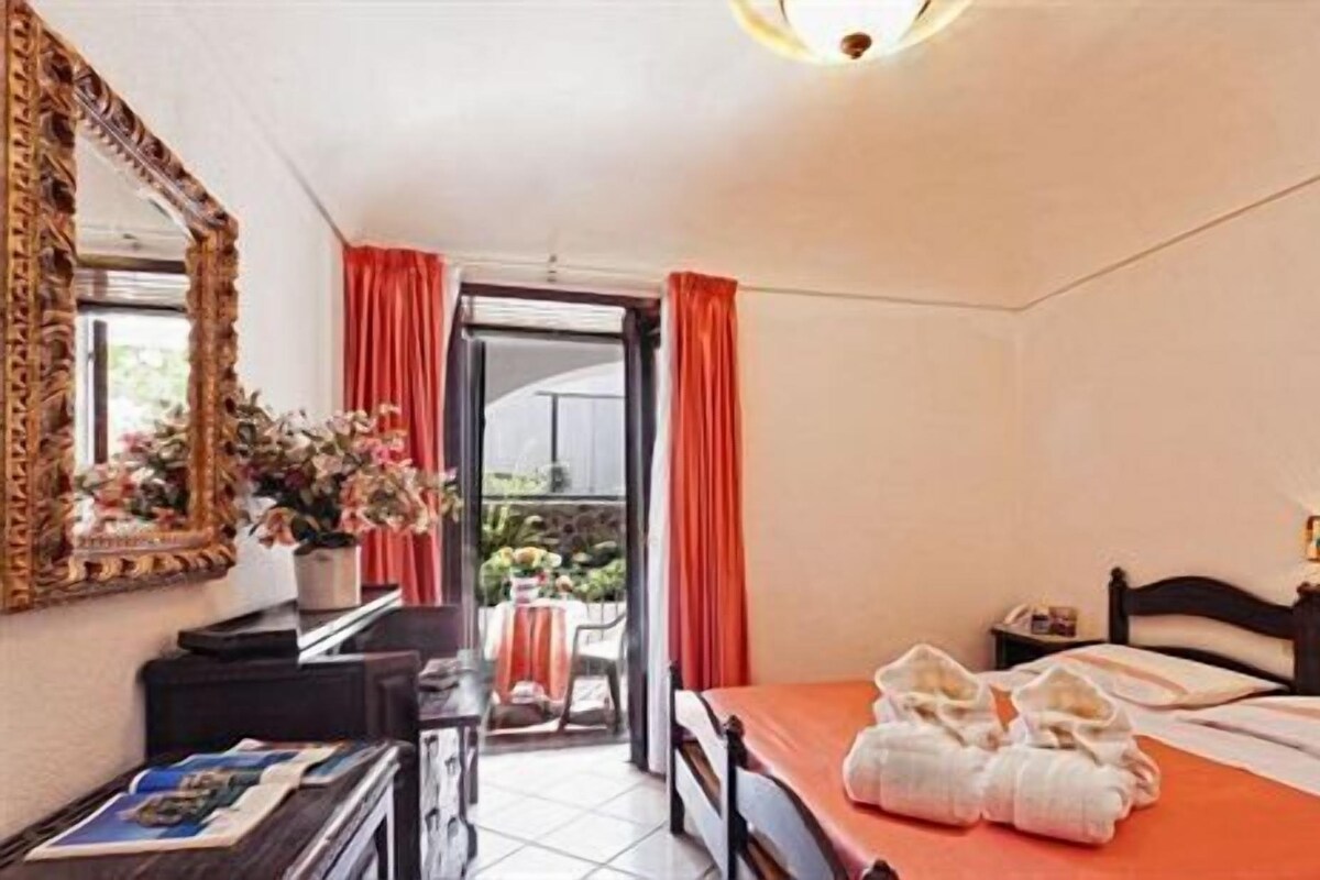 A hotel room with a bed, a desk, a mirror, and a view of a garden through a sliding door.