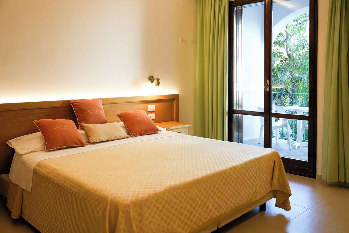 A hotel room with a bed, a nightstand, a window with curtains, and a view of the outdoors.