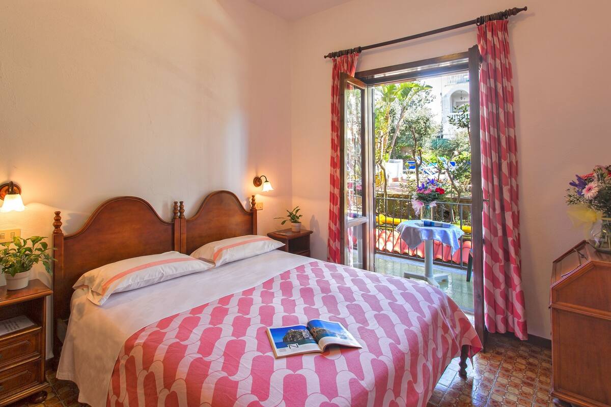 A neatly made bed with a patterned bedspread, a bedside table with a lamp, and a window with curtains.